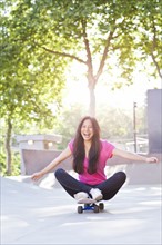 Cheerful young woman sitting on skateboard. Photo : Take A Pix Media