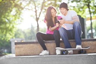 Young multi-racial couple sitting with skateboard, using mobile phone. Photo : Take A Pix Media