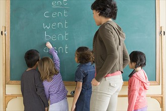 Children (6-7) writing on blackboard with teacher looking at them. Photo : Rob Lewine