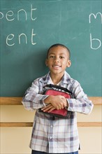 Smiling boy (6-7) holding lunch box against blackboard. Photo: Rob Lewine