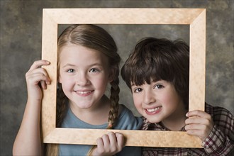 Portrait of girl (8-9) and boy (6-7) looking through frame, studio shot. Photo : Rob Lewine