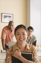 Young businesswoman posing for portrait with two colleagues in background. Photo : Rob Lewine