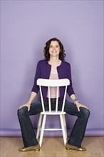 Portrait of smiling woman sitting on chair with purple background, studio shot. Photo : Rob Lewine
