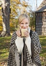 Portrait of smiling woman holding mug in front of cottage house in Autumn. Photo : Tetra Images