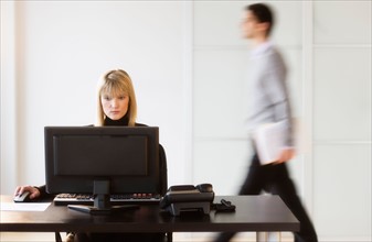 Businesswoman working on computer, businessman walking in background.
