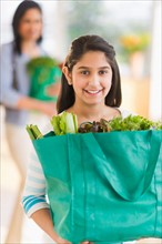 Daughter (12-13) holding bag of fresh vegetables, mother in background.