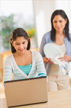 Girl using laptop (12-13) at home, mother in background.