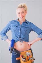 Portrait of young woman wearing tool belt and holding drill, studio shot. Photo : Daniel Grill