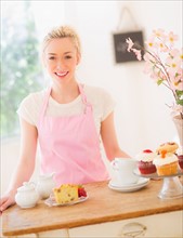 Smiling young woman in apron standing at table with cakes. Photo : Daniel Grill