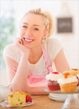 Smiling young woman in apron leaning on table with cakes. Photo : Daniel Grill