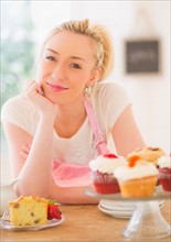 Smiling young woman in apron leaning on table with cakes. Photo : Daniel Grill