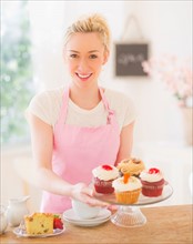 Smiling young woman in apron holding cake stand . Photo : Daniel Grill