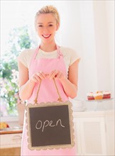 Smiling young woman in apron holding open sign. Photo : Daniel Grill