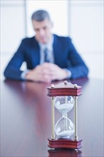 Hourglass on desk, businessman sitting on background. Photo : Daniel Grill
