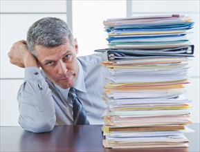 Businessman sitting next to large stack of documents. Photo : Daniel Grill