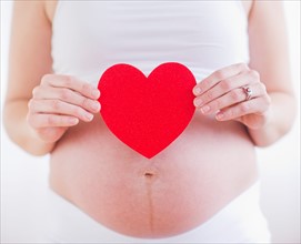 Pregnant woman holding red paper heart on her belly. Photo : Daniel Grill