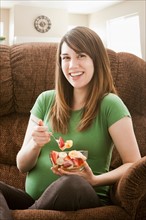 Portrait of pregnant woman sitting on sofa and eating salad. Photo : Mike Kemp