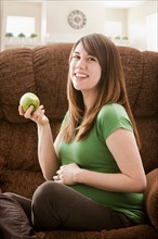 Portrait of pregnant woman sitting on sofa and eating apple. Photo : Mike Kemp