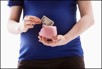 Studio Shot of mid section of pregnant woman putting money into Piggy Bank. Photo : Mike Kemp