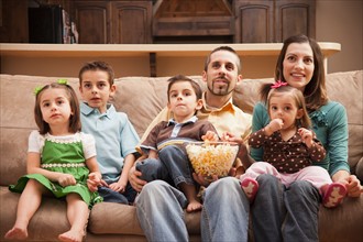 Front view of family with children (18-23 months, 4-5, 6-7, 8-9) sitting on couch watching tv and