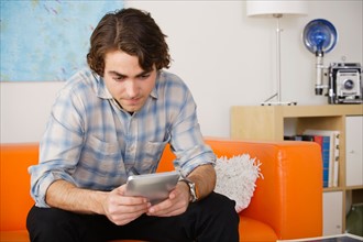 Young man sitting on sofa and using digital tablet. Photo : Rob Lewine