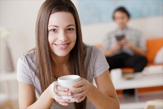 Portrait of young woman holding mug, man in background. Photo : Rob Lewine