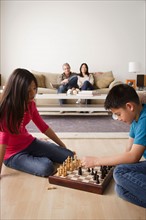 Siblings playing chess game while parents watching from sofa. Photo : Rob Lewine
