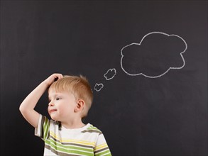 Cute toddler boy (2-3) scratching his head against blackboard with thought bubble written in chalk.