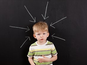 Cute toddler boy (2-3) standing against blackboard with arrows written in chalk pointing at him.