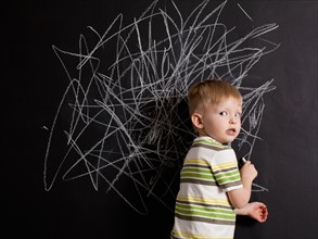 Cute toddler boy (2-3) standing against blackboard with daub written in chalk. Photo : Jessica