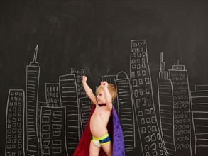 Cute toddler boy (2-3) standing against blackboard with city skyline drawn on it. Photo : Jessica