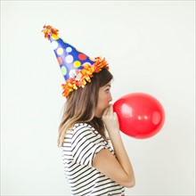 Studio shot of young woman wearing party hat and blowing balloon. Photo : Jessica Peterson