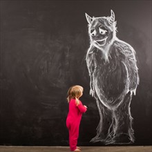 Portrait of baby girl (18-23 months) in front of huge monster drawn on blackboard. Photo : Jessica