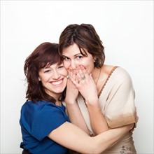 Studio Shot portrait of young women embracing, covering mouth with hand. Photo : Jessica Peterson