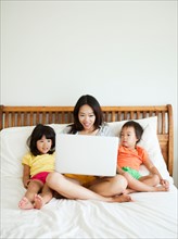 Mother with daughters (2-3, 4-5) sitting on bed, using laptop. Photo : Jessica Peterson