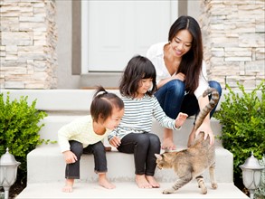 Mother sitting on doorsteps with two daughters (2-3, 4-5) and playing with cat. Photo : Jessica