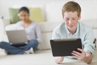Boy (16-17) lying on floor using tablet pc, girl (14-15) with laptop in background.