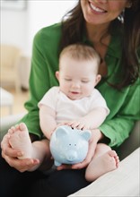 Mother holding baby daughter (6-11 months) and piggy bank. 
Photo : Jamie Grill