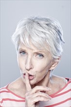 Portrait of senior woman with finger on her lips, studio shot. 
Photo : Rob Lewine
