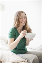 Happy young woman sitting on bed with cornflake bowl. 
Photo : Jessica Peterson