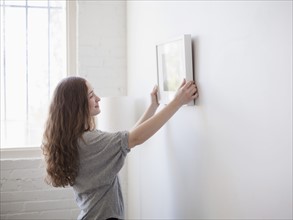 Young woman hanging picture on freshly painted wall. 
Photo : Jessica Peterson