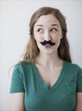 Studio shot of happy young woman with fake moustache. 
Photo: Jessica Peterson