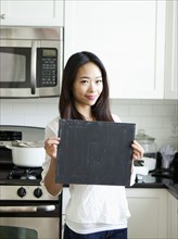 Portrait of smiling young woman holding blackboard in kitchen. Photo : Jessica Peterson