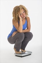 Portrait of young woman crouching on weight scale, studio shot.