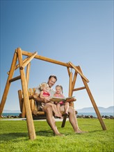 Father with daughters (2-3, 4-5) on swing reading book.