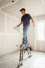 Drywall worker on stilts walking across unfinished room. Photo: Mike Kemp