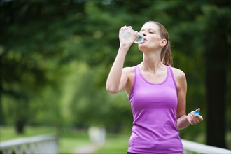 Young woman walking over a bridge and drinking a bottle of water after jogging
