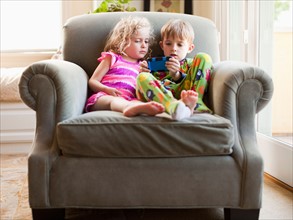 Girl and boy sitting on armchair and playing video game. Photo : Jessica Peterson