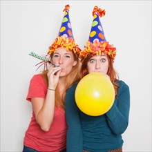 Studio Shot, Two women wearing party hats and one blowing up yellow balloon. Photo : Jessica