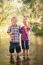 Little boy (6-7) and little girl (4-5) standing together in small steam holding wooden stick fishing poles